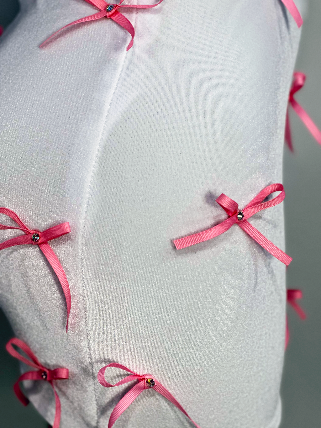 A two-piece skirt set with a white fitted tank top and a white high-waisted mini skirt, both adorned with pink ribbon bows and rhinestone accents by Bellz Couture in Detroit. shown on mannequin against a plain wall.