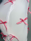 A two-piece skirt set with a white fitted tank top and a white high-waisted mini skirt, both adorned with pink ribbon bows and rhinestone accents by Bellz Couture in Detroit. shown on mannequin against a plain wall.